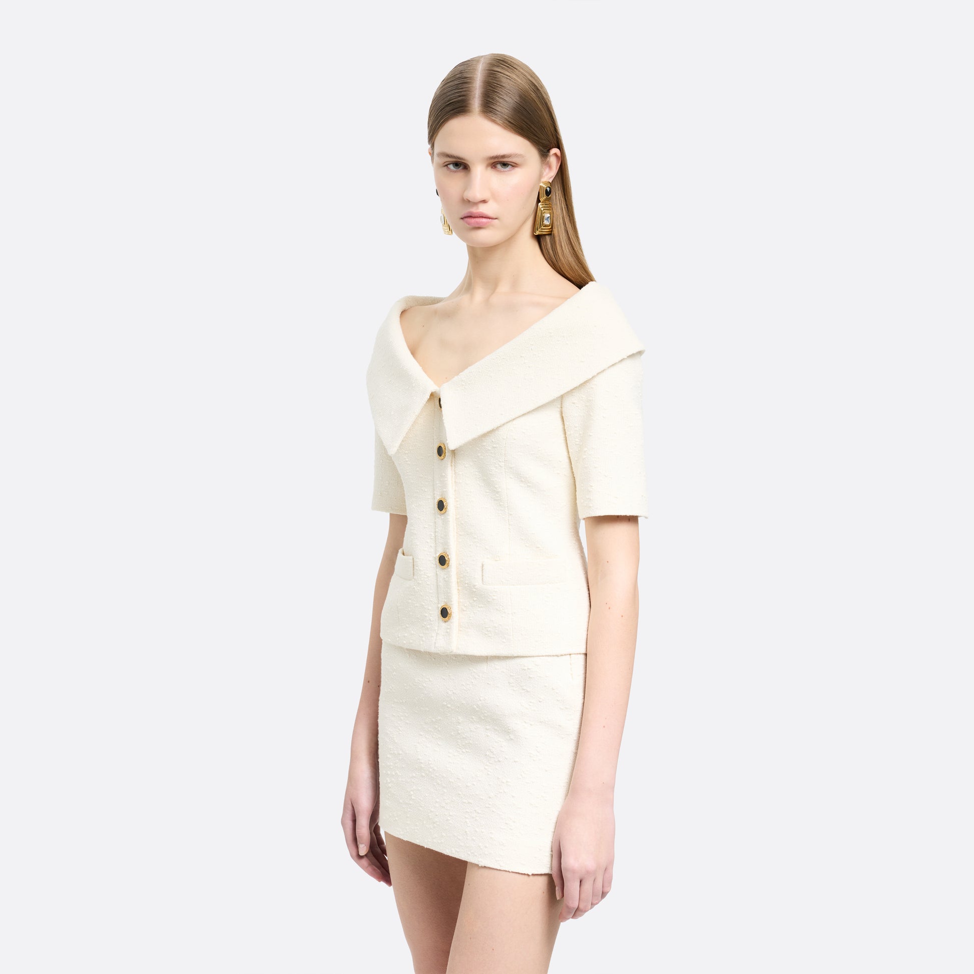 Ivory tweed bouclé off-shoulder jacket with a wide folded neckline and short sleeves. Finished with black and gold-tone buttons and welt pockets.