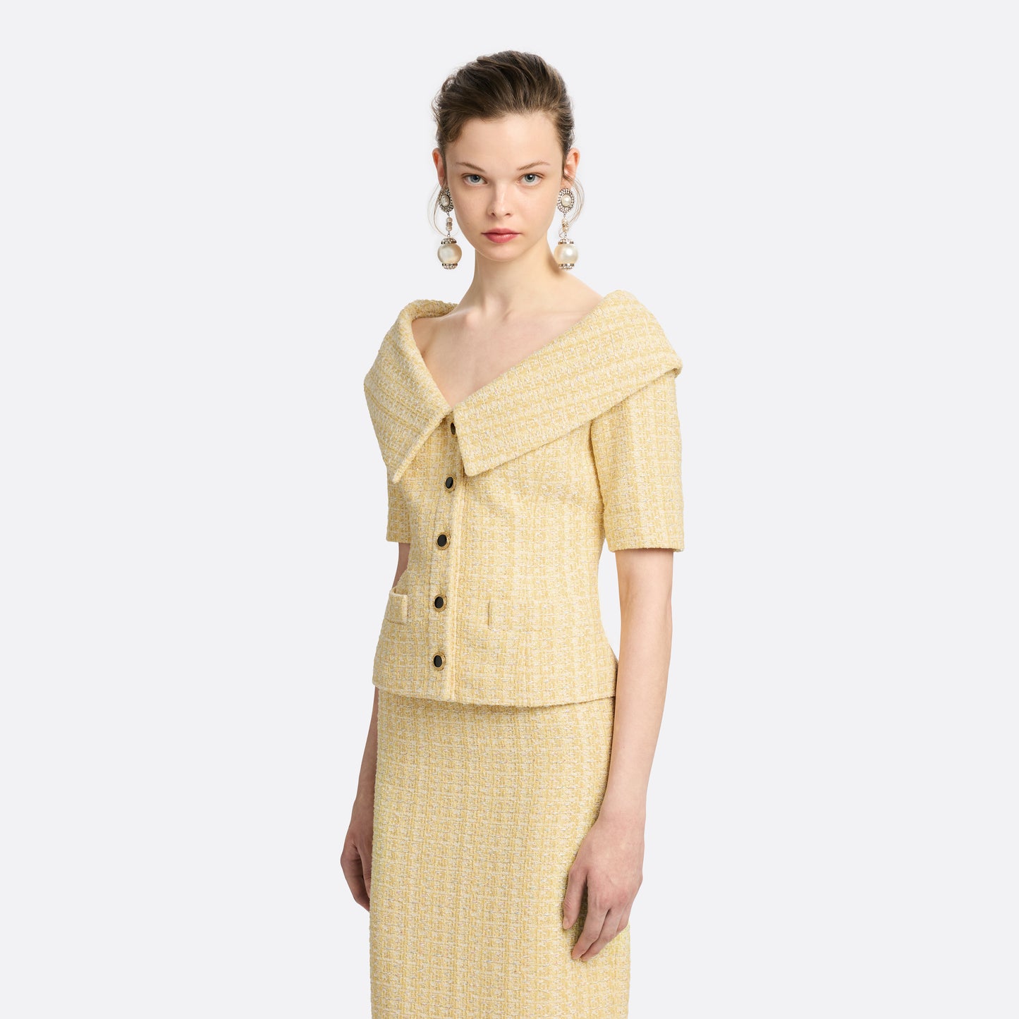 Pale yellow sequin tweed off-shoulder jacket with a wide folded neckline and short sleeves. Finished with black and gold-tone buttons and welt pockets.