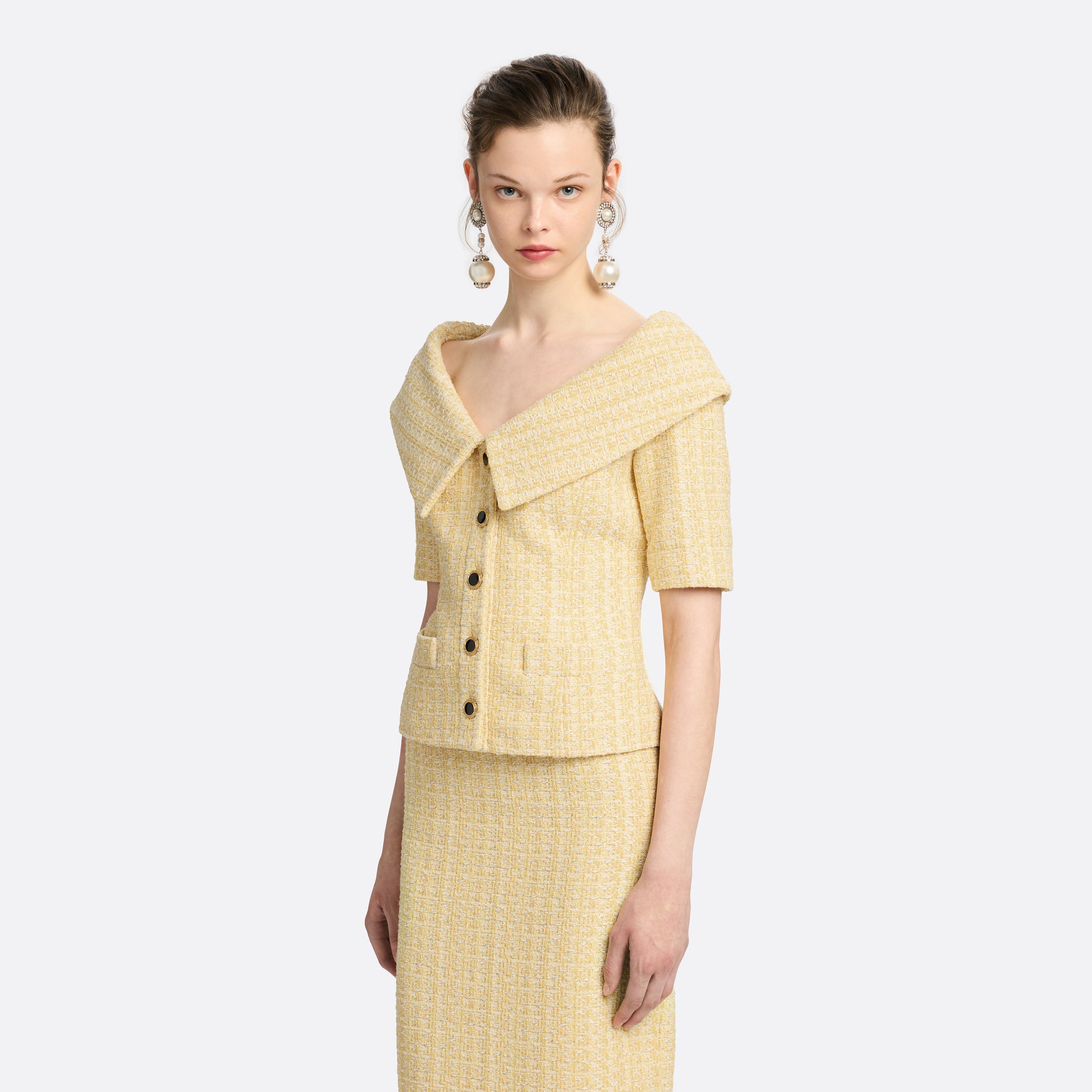 Pale yellow sequin tweed off-shoulder jacket with a wide folded neckline and short sleeves. Finished with black and gold-tone buttons and welt pockets.