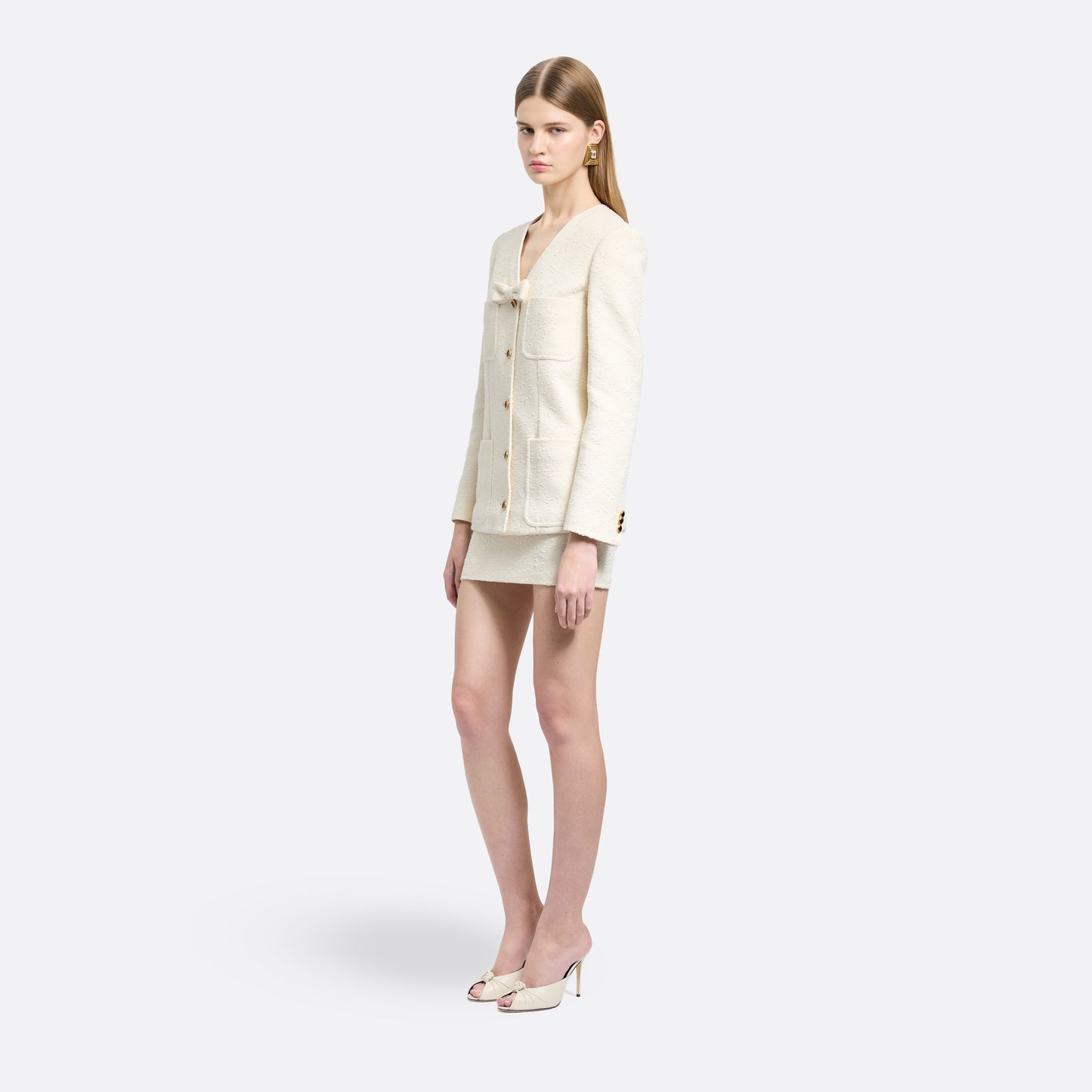 Ivory tweed bouclé jacket cut to a longline silhouette with a V-neckline and patch pockets. Finished with black and gold-tone buttons and a bow detail.