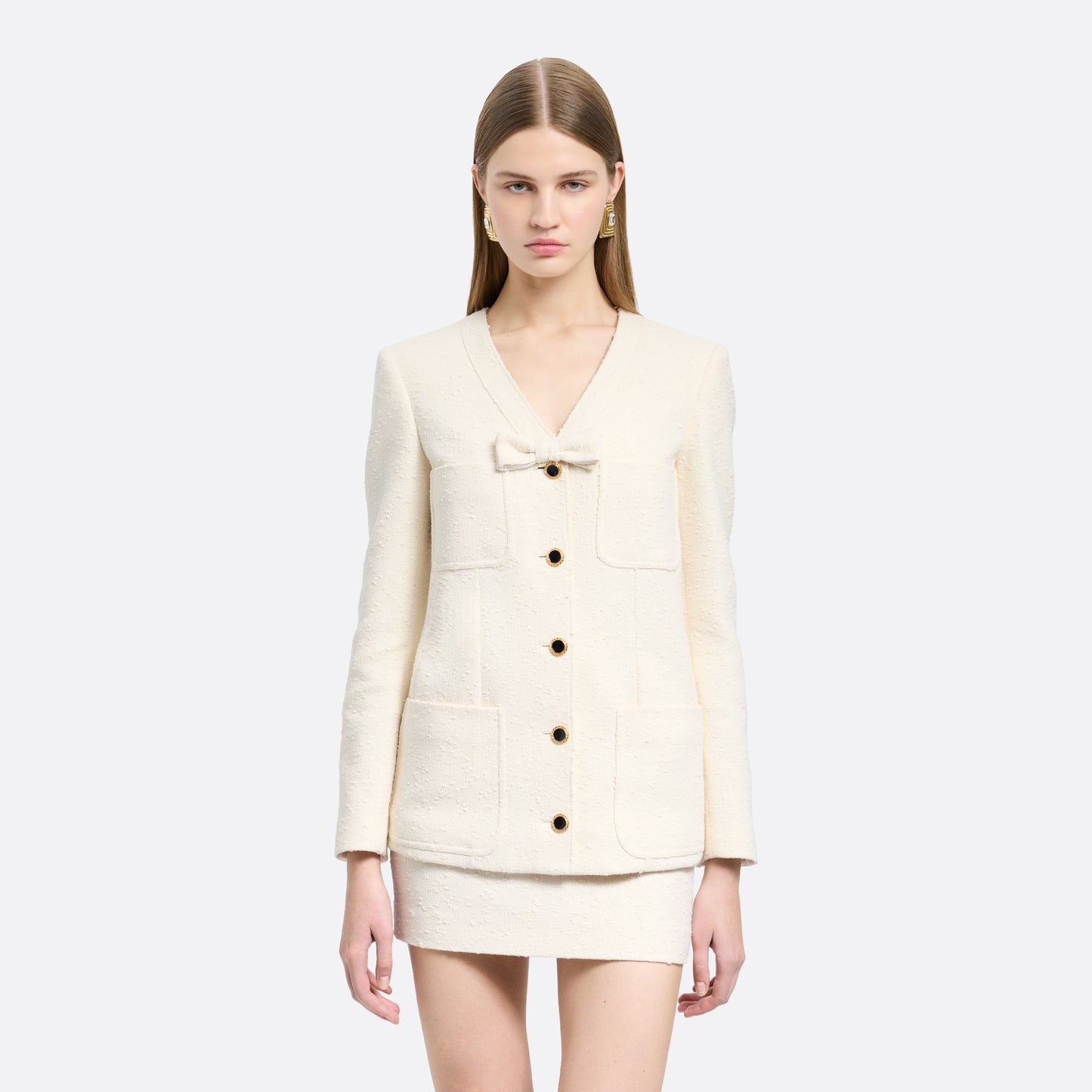 Ivory tweed bouclé jacket cut to a longline silhouette with a V-neckline and patch pockets. Finished with black and gold-tone buttons and a bow detail.