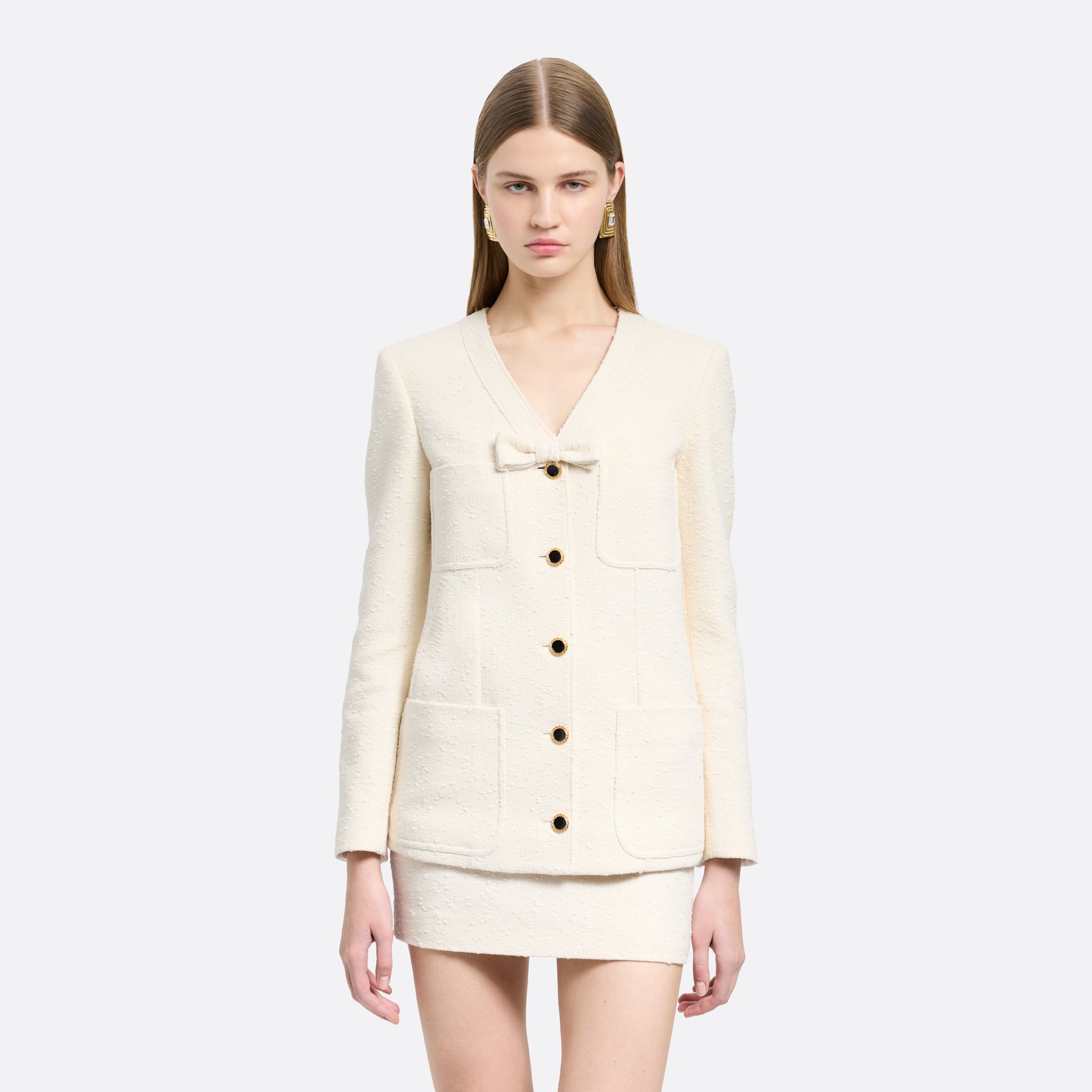 Ivory tweed bouclé jacket cut to a longline silhouette with a V-neckline and patch pockets. Finished with black and gold-tone buttons and a bow detail.